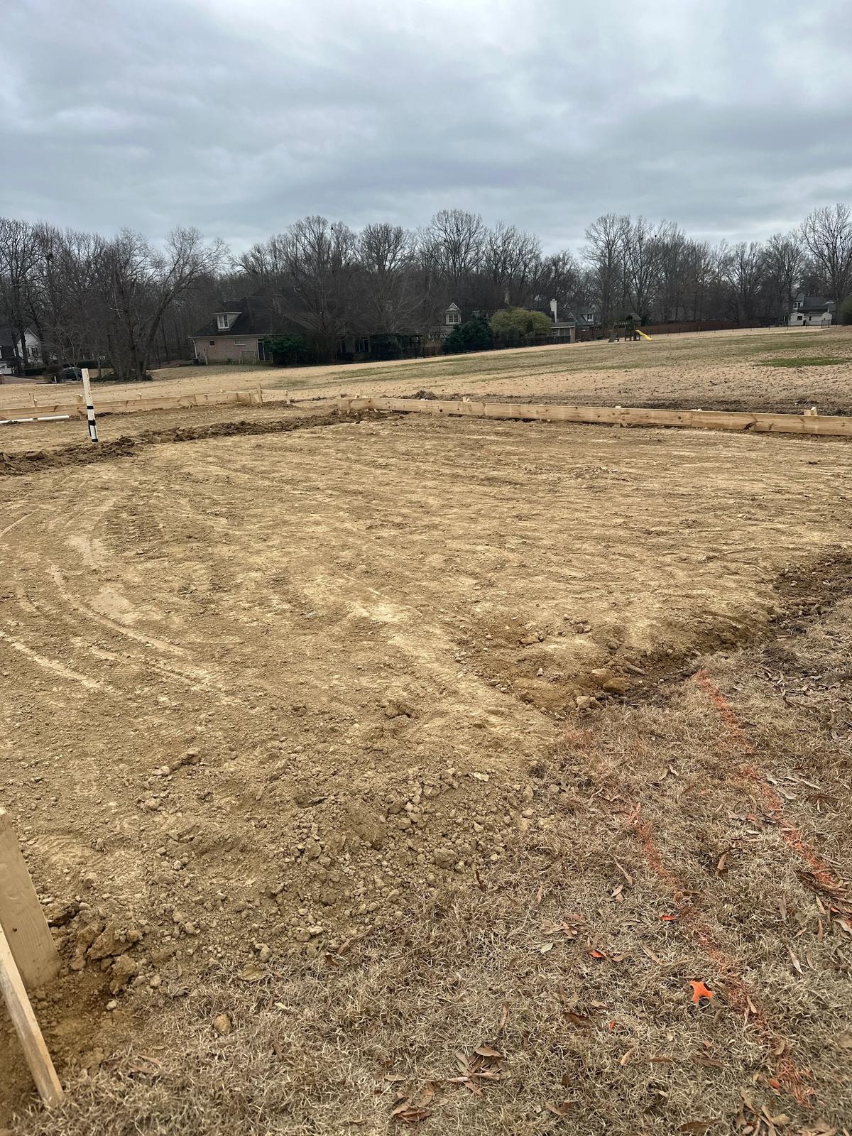Cleared building site with wood stakes and string lines marking out a foundation, houses visible in the background.