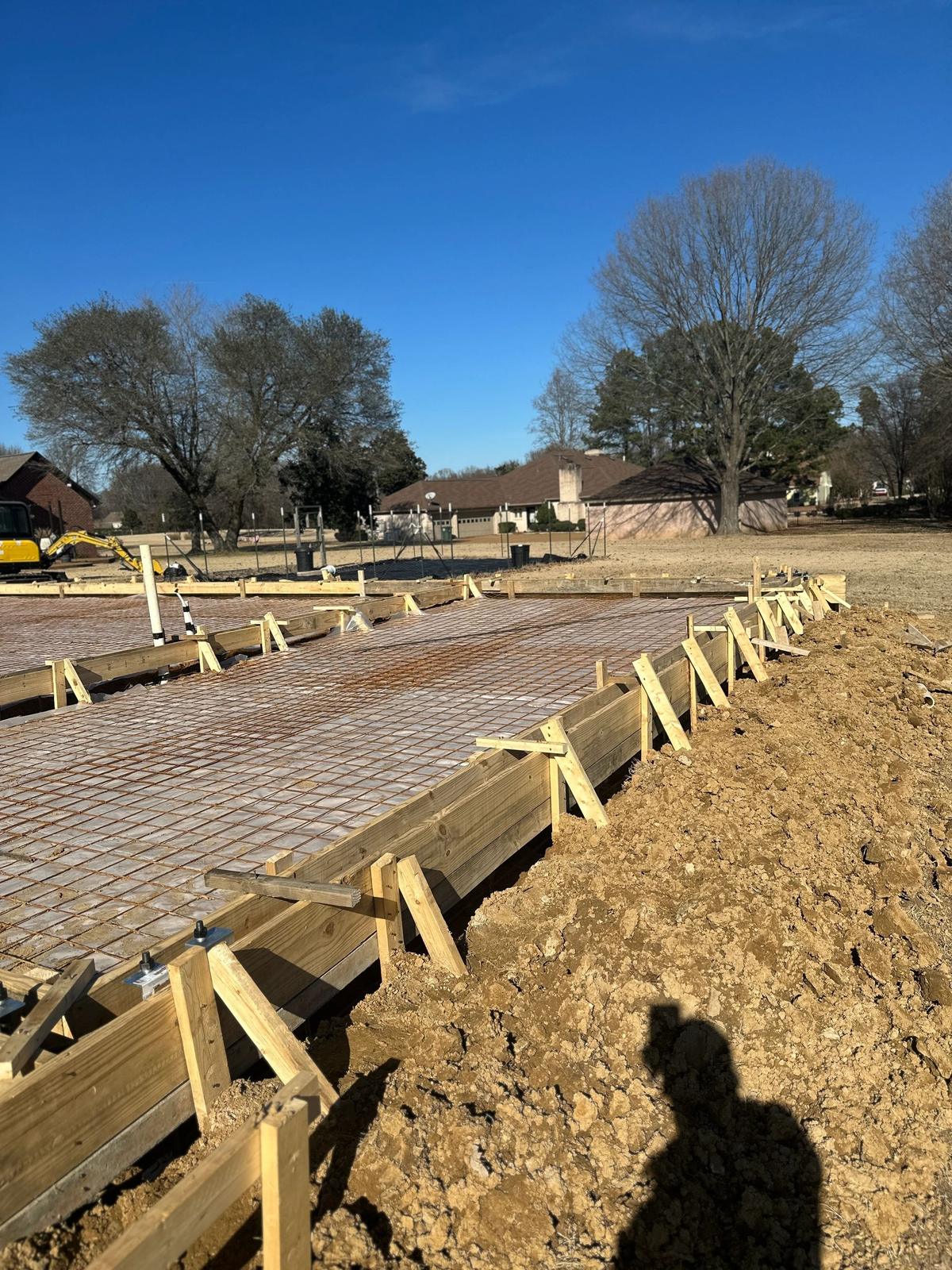 Concrete slab foundation with a rebar grid and wood forms, a mini excavator and house visible behind it.