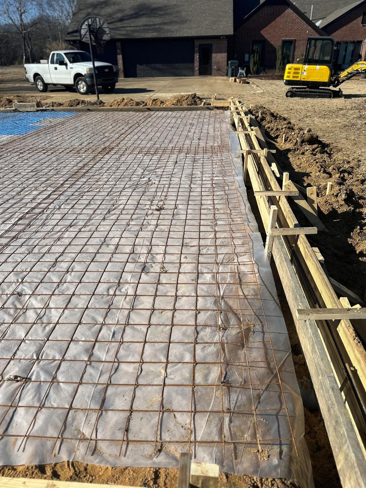 Rebar and vapor-barrier slab foundation with wood forms, a pickup truck and mini excavator near a brick house.
