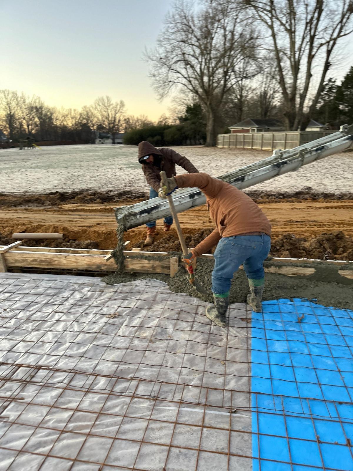 Two workers in cold-weather gear pour concrete from a chute onto a rebar-reinforced slab at dusk.