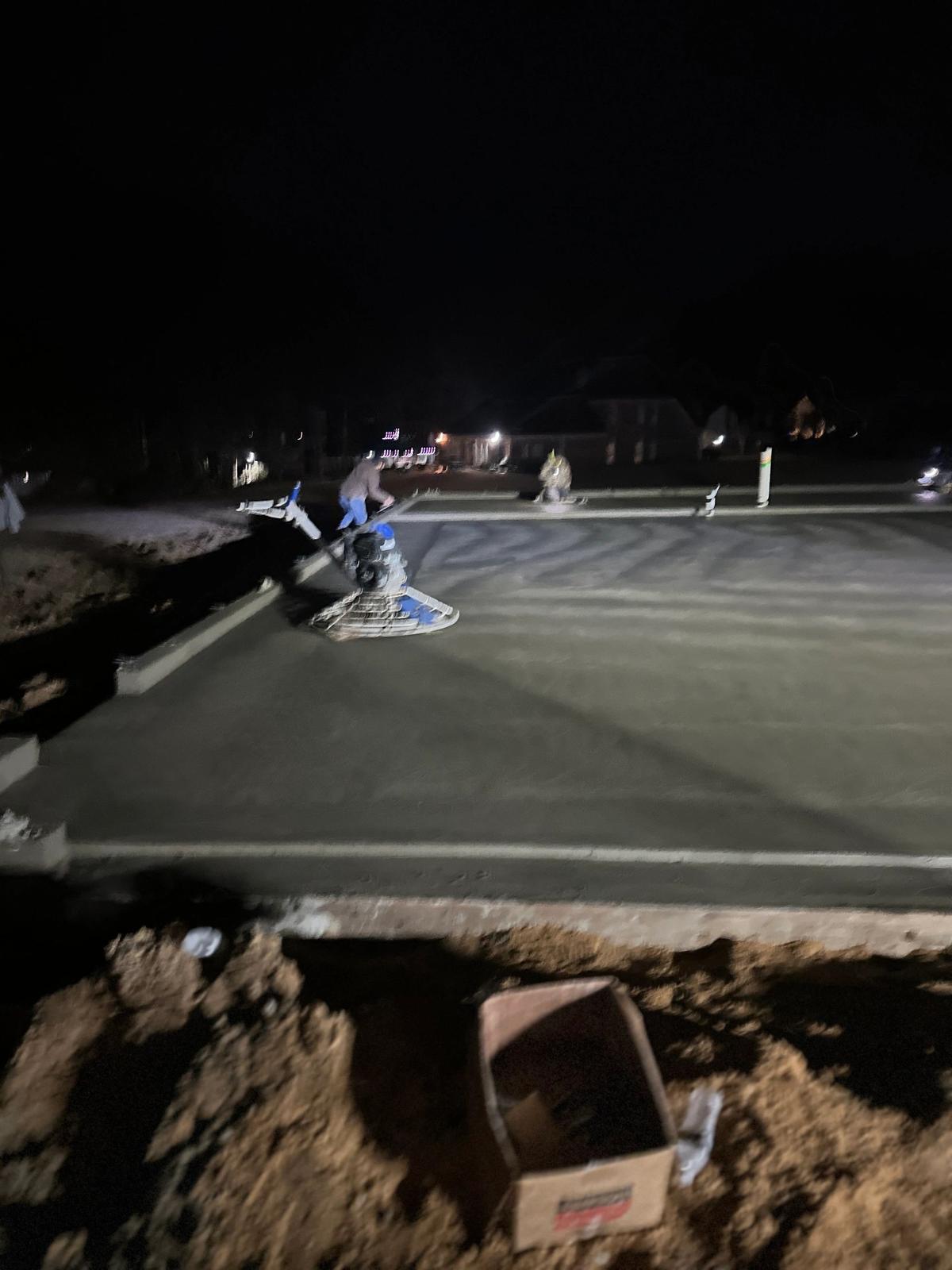 Blurry nighttime photo of a power trowel finishing a concrete slab; the image is dark and out of focus.