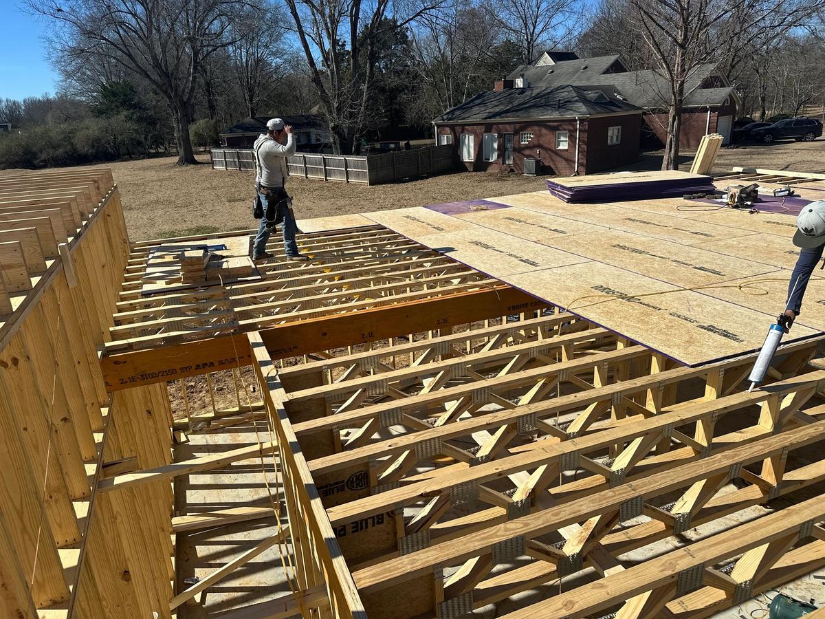 Worker on a roof deck installing plywood sheathing over wood floor trusses, with a brick house visible behind.