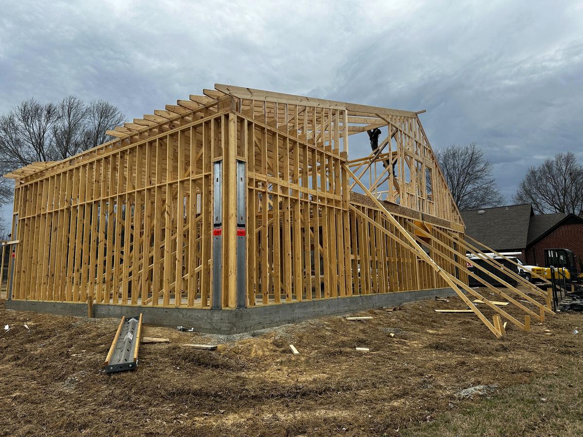 Large wood-framed building under construction with exposed wall studs and roof trusses, a worker visible near the peak.