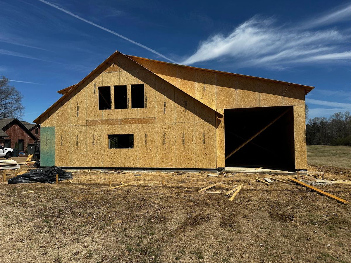 Large OSB-sheathed building with a dark open garage doorway and cut window openings, under a blue sky.
