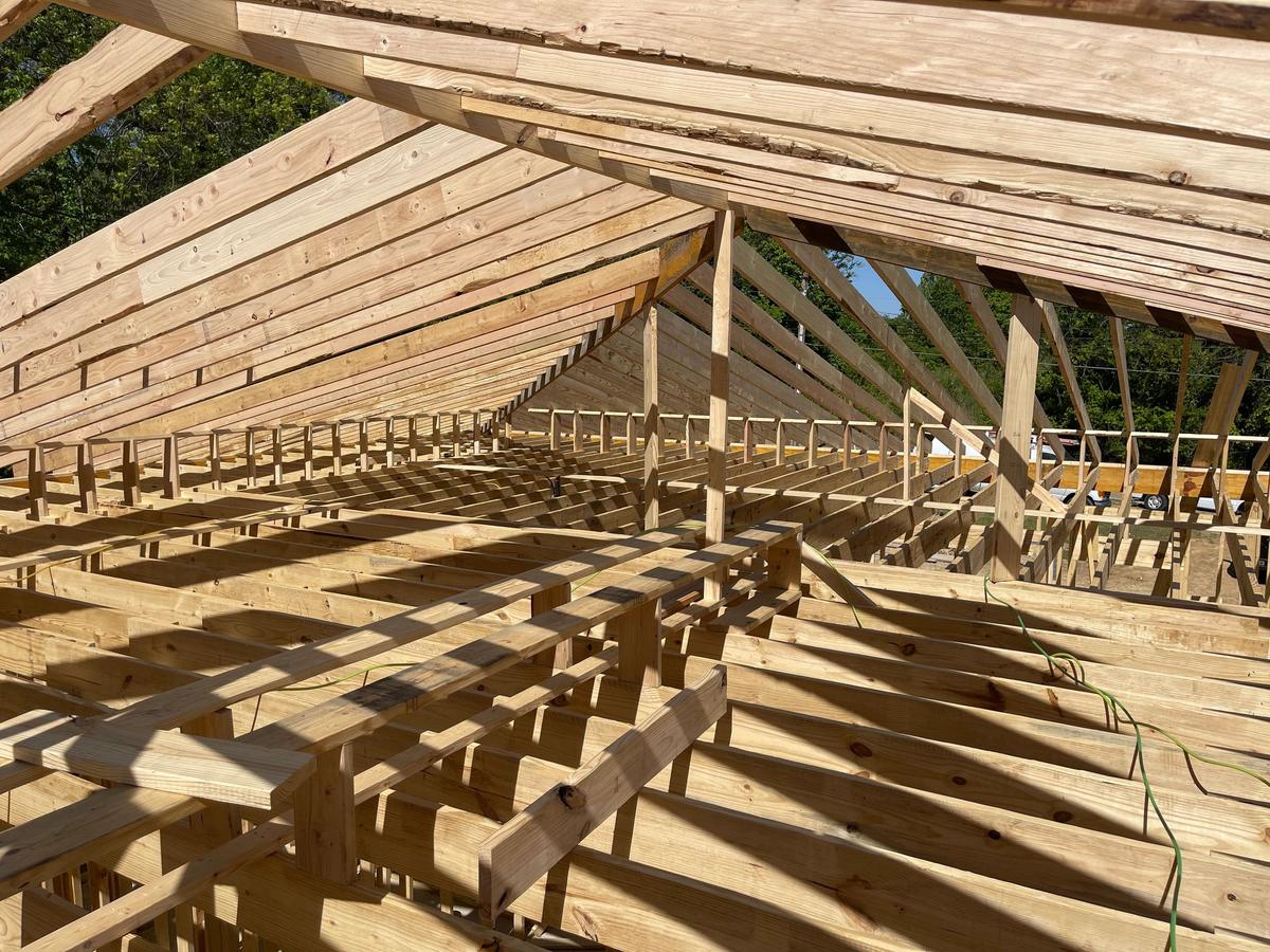 Close-up view down a row of exposed wood roof rafters and trusses on a large structure, sky visible through gaps.