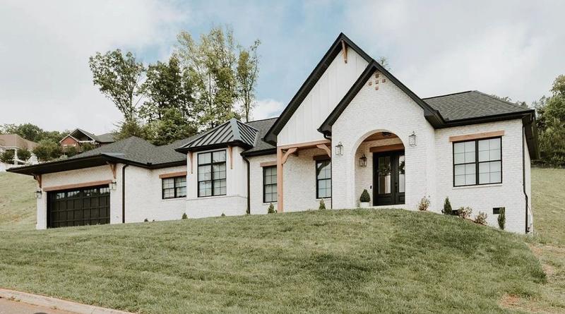 Finished single-story house with white brick, board-and-batten gables, a black roof and door, set on a grassy slope.