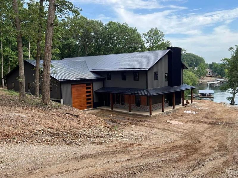 Finished dark-toned modern house with a wood accent garage door and metal roof, near a lake with a boat dock.