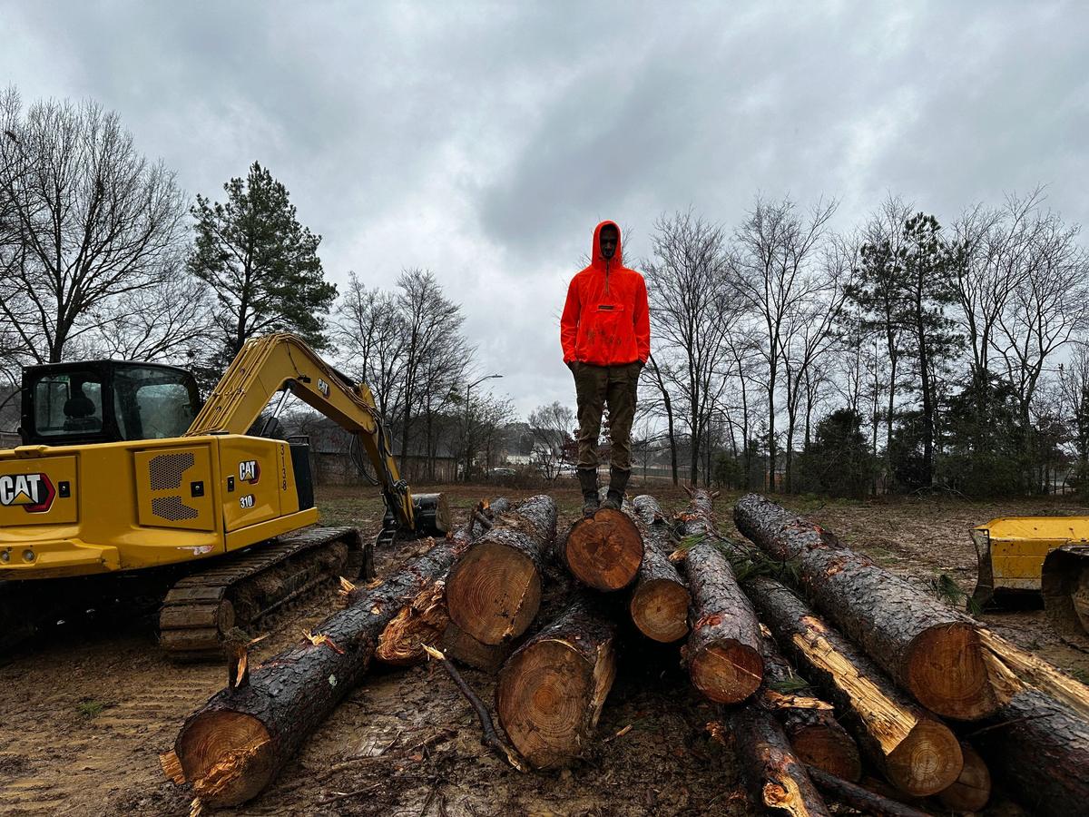 Man in an orange jacket stands on a pile of cut logs next to a yellow excavator on a cleared, muddy site.